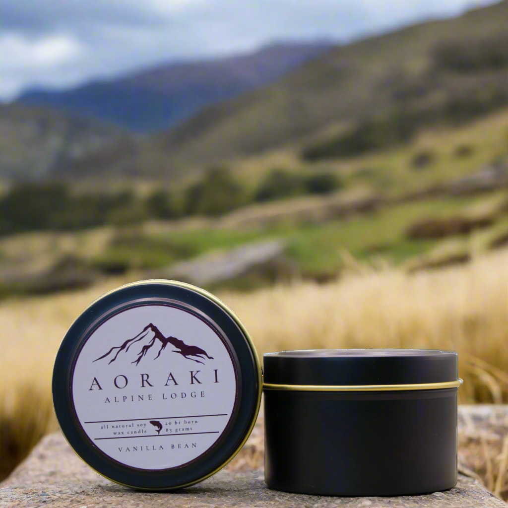 A photo of a Black Candle Tin with a customised logo, taken in a mountainous area with tussock in the background. It is an example of what the Wild Essentials Custom Candles could look like when produced as Promotional Merchandise for a Corporate Client, in this case Aoraki Alpine Lodge.