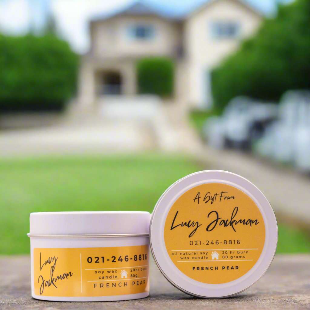 Photo of a 85g Real Estate Custom Candle from Wild Essentials. White candle tin, yellow label. In the background is a home.