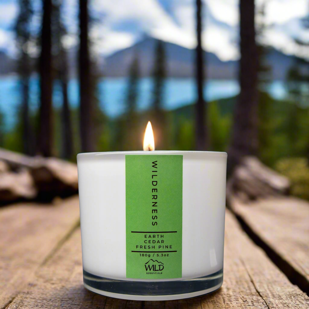 Photo of the Wilderness Luxury Candle from Wild Essentials. The candle is resting on some worn wood. Possibly an old, worn, outdoor table. In the background are pine trees, water, and some mountains.