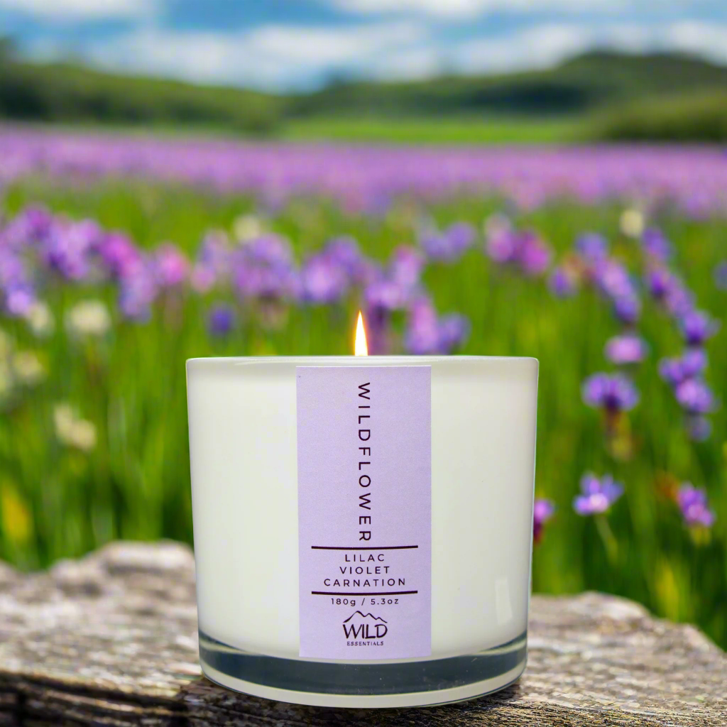 Photo of the Wildflower Luxury Candle from Wild Essentials. The candle is positioned on a rock in a meadow. In the background is a field of wildflowers and rolling hills.