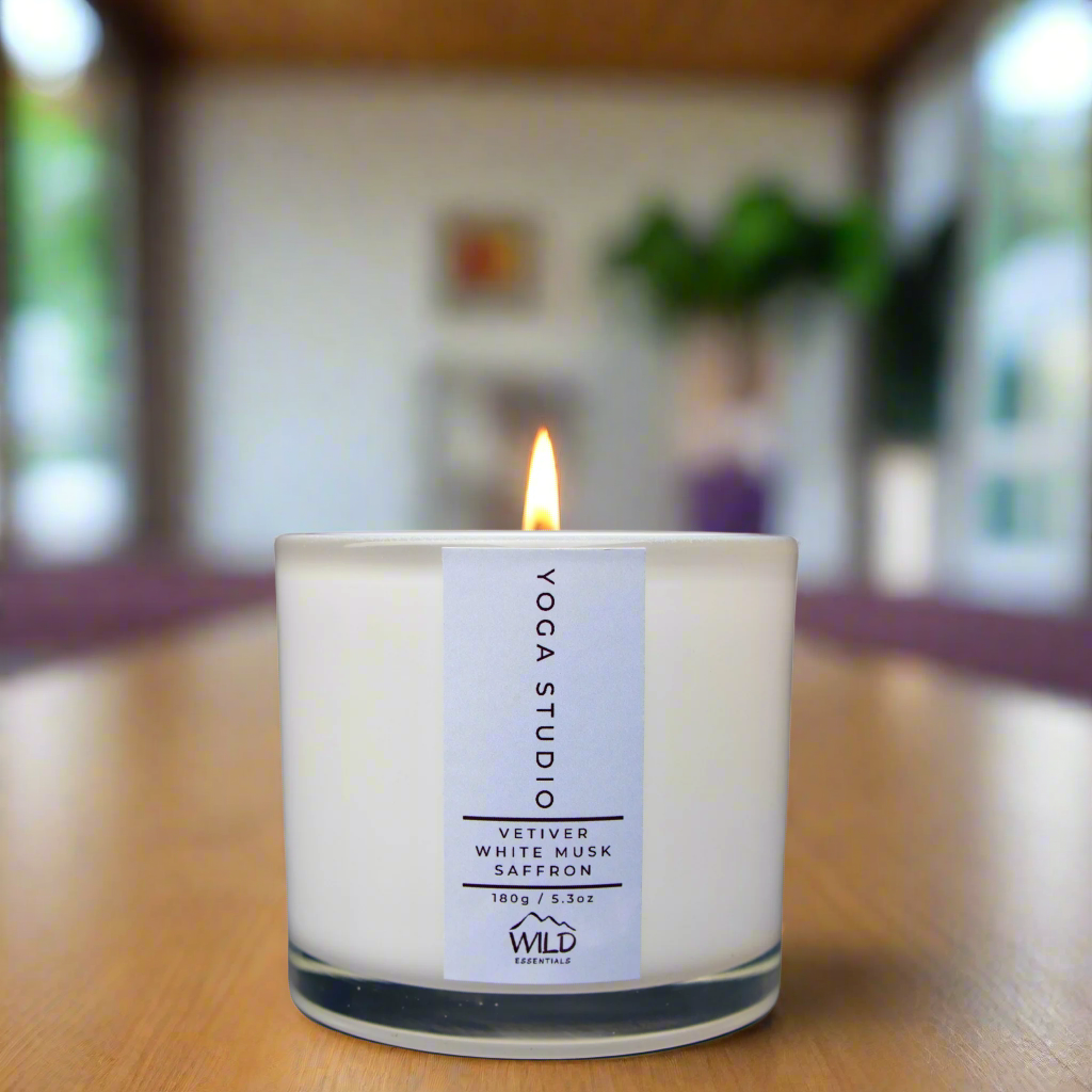 A photo of the Yoga Studio Luxury Candle from Wild Essentials. The background is blurry but yoga mats can be seen in the background and also some green plants, both inside and out.