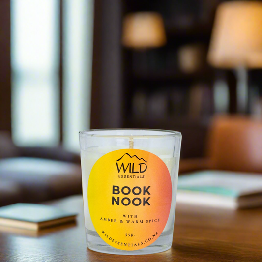 Photo of the Book Nook Mini Candle made by Wild Essentials. The candle is on a wooden table in a study with books , lamps, and a book shelf nearby.