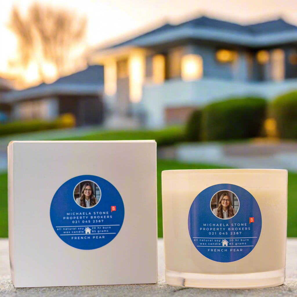 Photo of the 200g Luxury Real Estate Candle with box from Wild Essentials. A home can be seen in the background.
